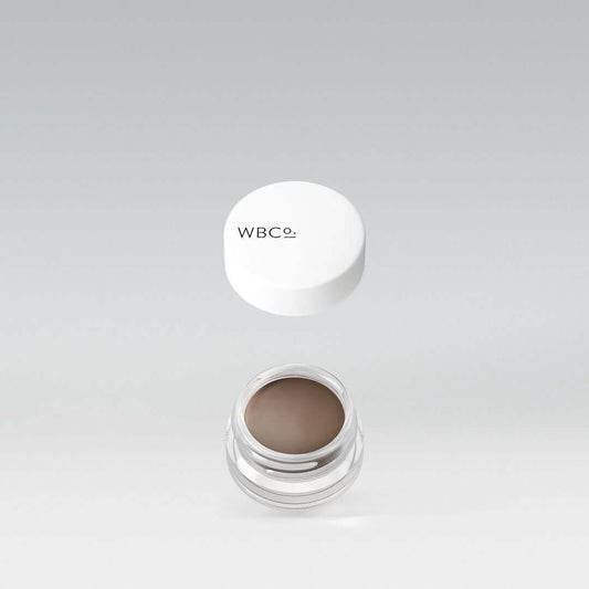 A jar of eyebrow pomade with a white lid, labeled 'WBCo', on a light background.


